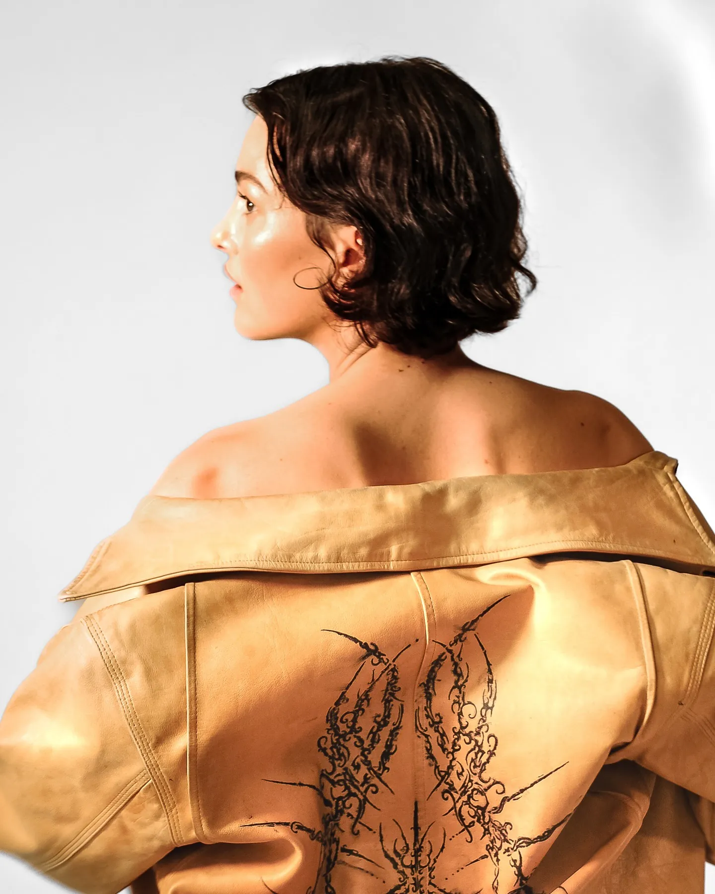 Model van achteren in een cognackleurige leren jas met zwarte print, glowy huid in profiel