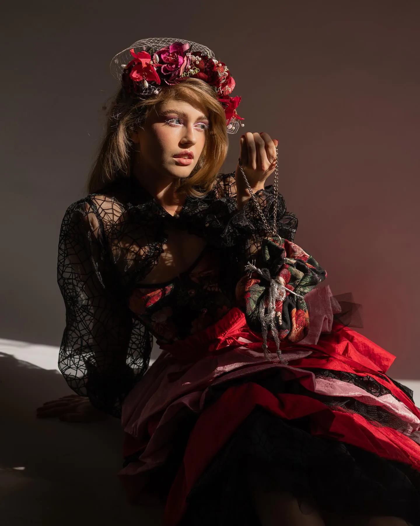 Model met bloemenhoed en voile in zijlicht, kanten blouse met spinnenwebmotief en gelaagde rode rokken