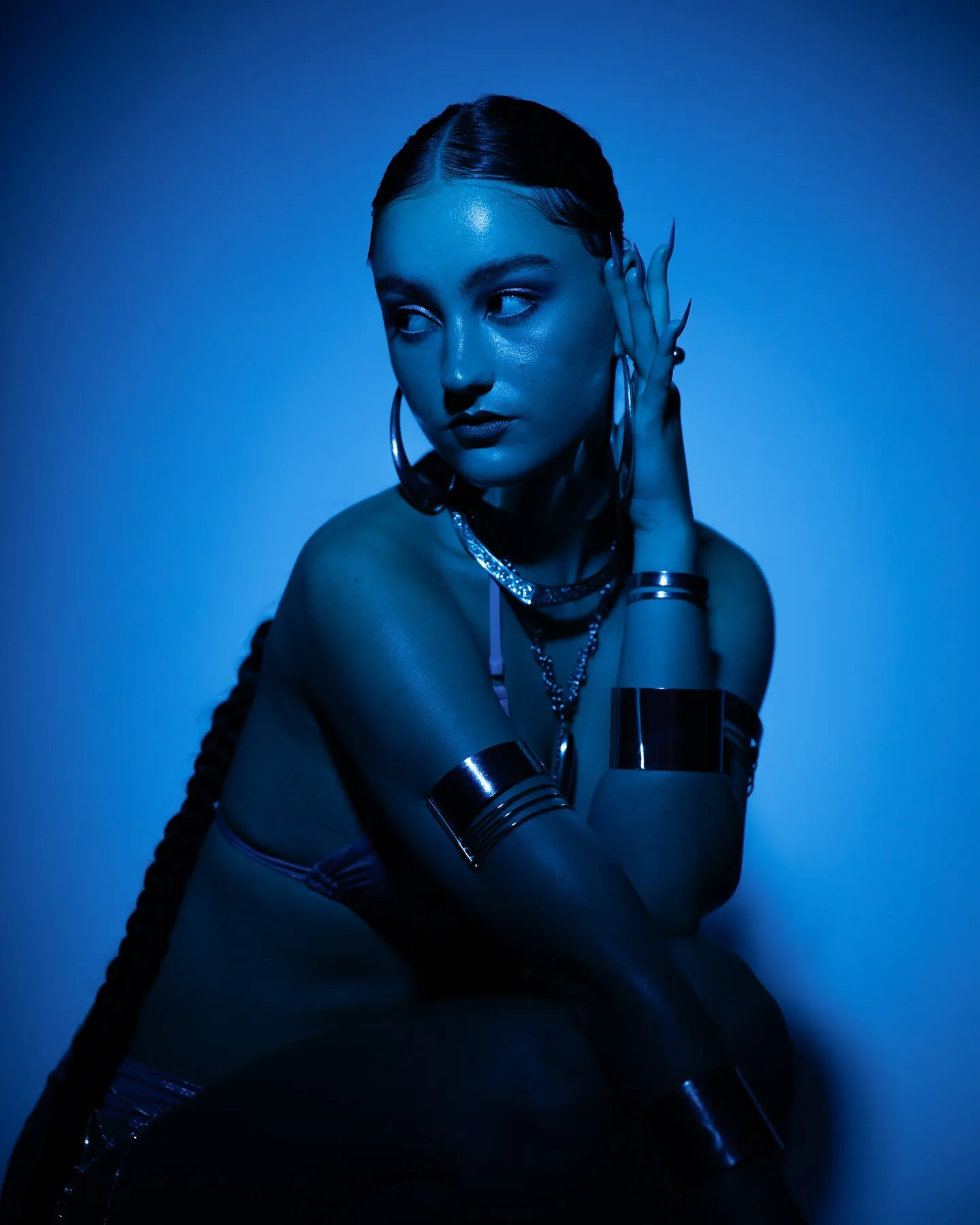 Gehurkt model in blauw licht kijkt over haar schouder, lange vlecht en zilveren armbanden