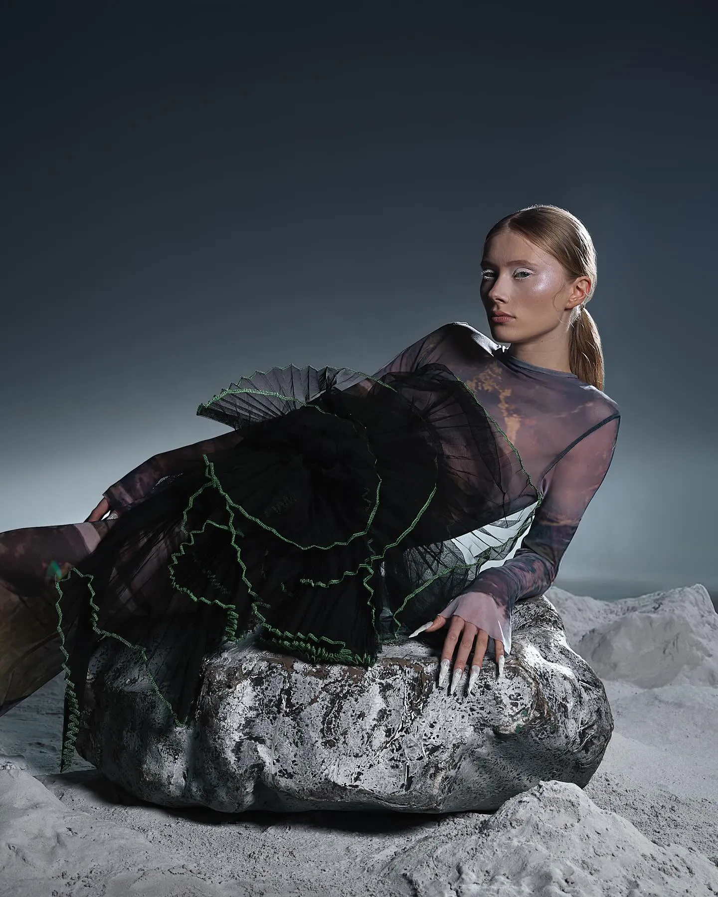 Model ligt op een zilveren rots in een grijs maanlandschap, witte wimpers en glanzende jukbeenderen