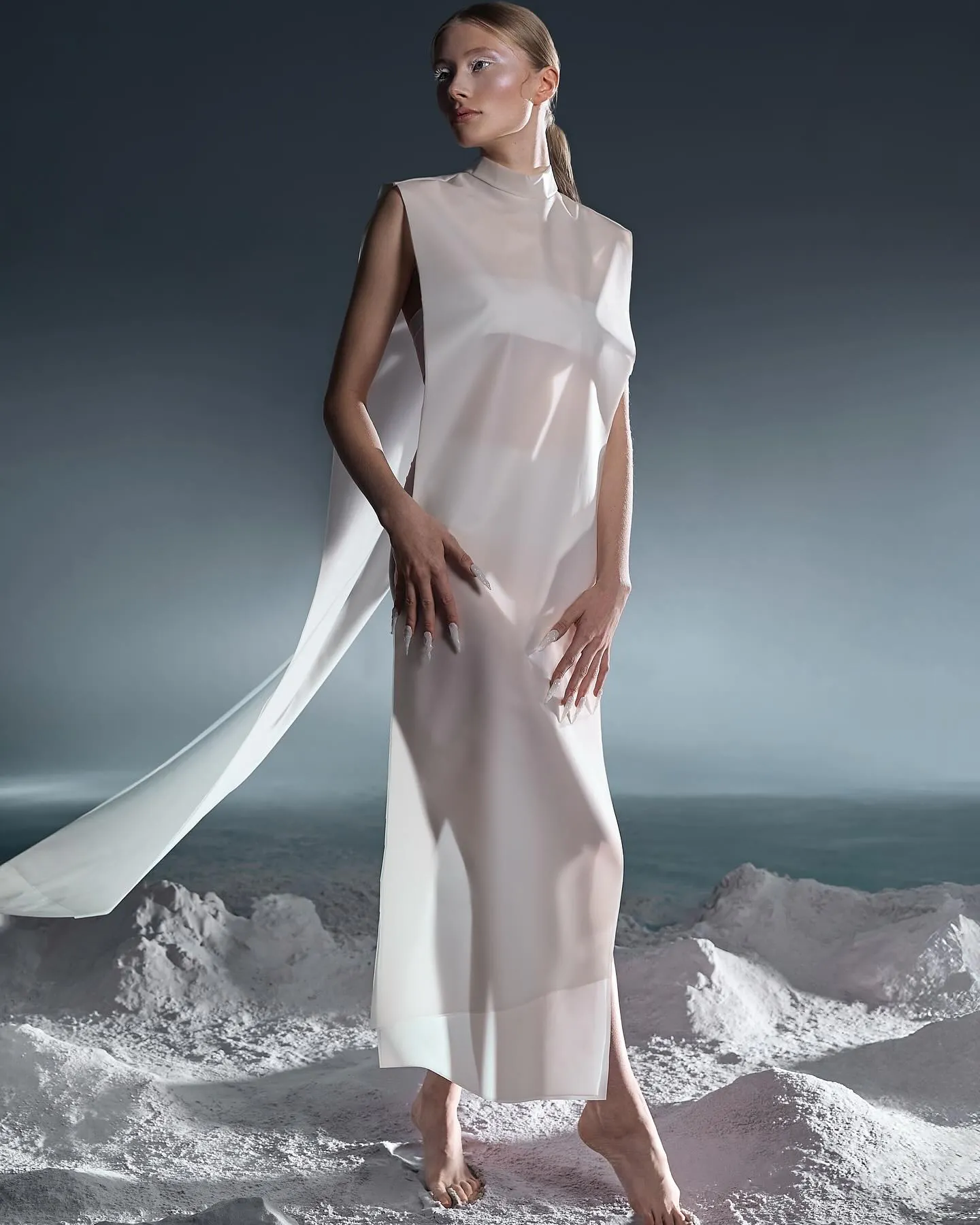 Model in een doorschijnende witte jurk met lange sleep staat op witte zandheuvels