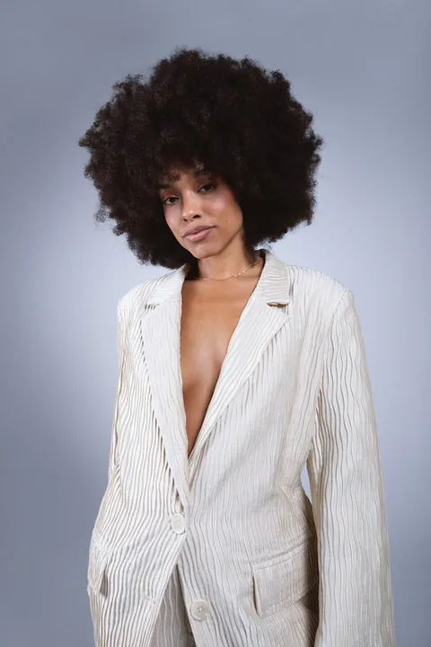 Vrouw met een grote afro en natuurlijke make-up in een crèmekleurige geplisseerde blazer, tegen een grijsblauwe achtergrond
